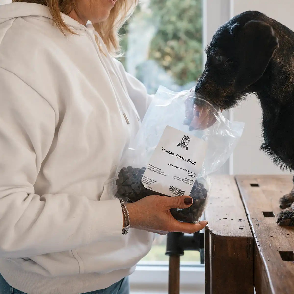 Dackel sitzt auf einem Tisch und wird mit den Trainee Treats gefüttert und man sieht die Packung und der Hund nimmt eigenständig seinen Kopf dort rein um an die Leckerlis zu kommen. 