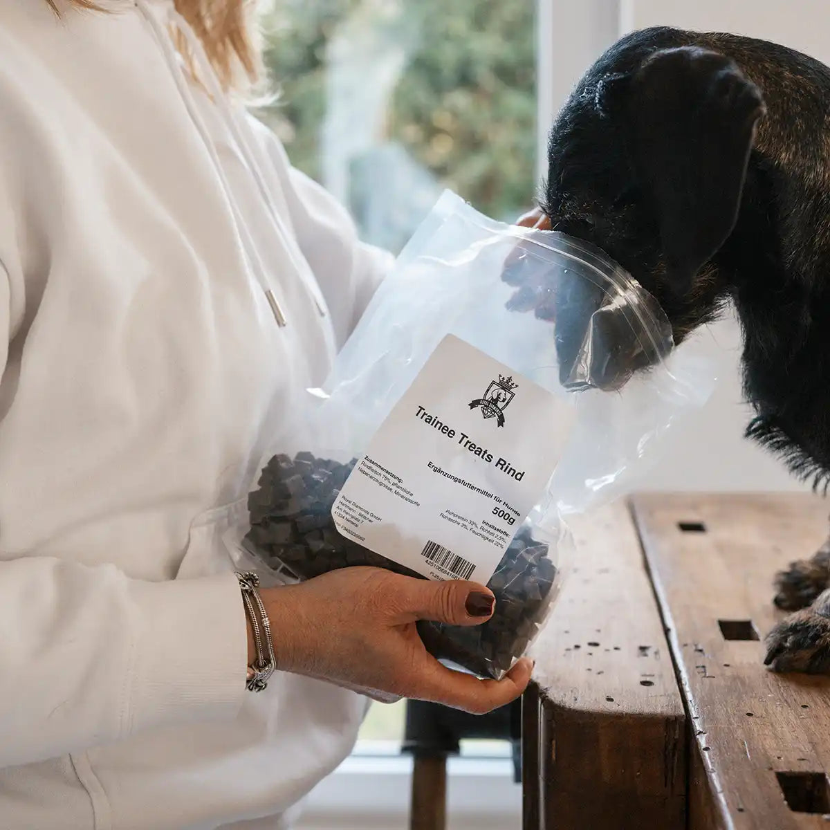 Dackel sitzt auf einem Tisch und wird mit den Trainee Treats gefüttert und man sieht die Packung und der Hund nimmt eigenständig seinen Kopf dort rein um an die Leckerlis zu kommen. 