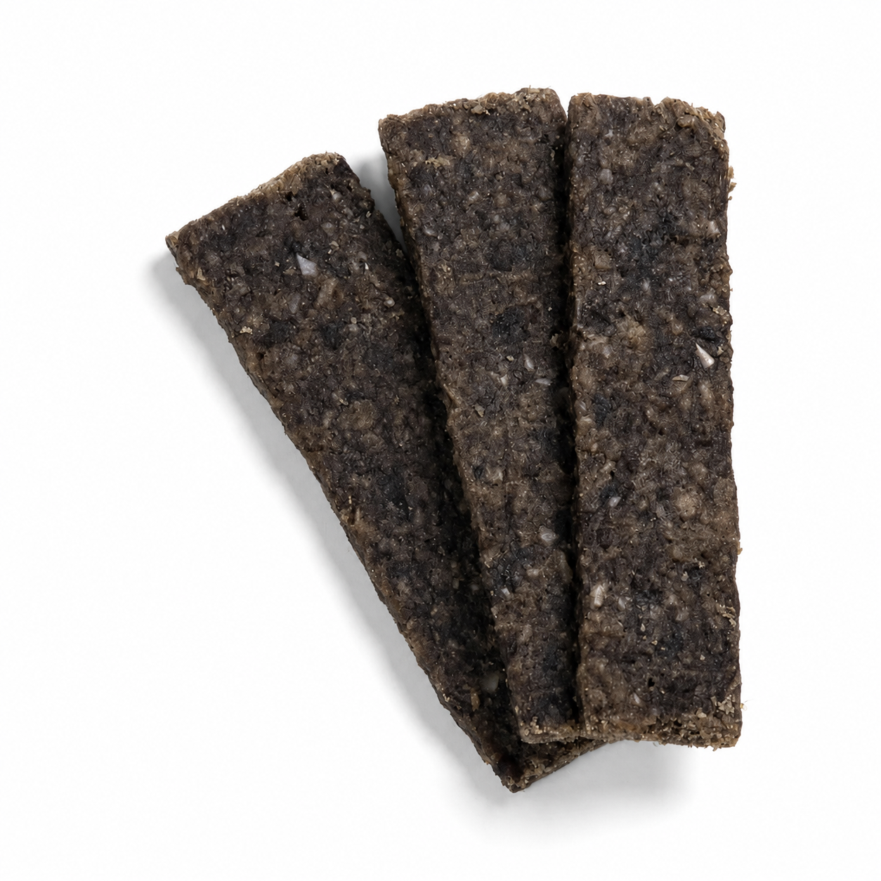 Das Bild zeigt drei dunkelbraune Hundesnack-Streifen in rechteckiger Form, die leicht fächerförmig und überlappend auf einem sauberen weißen Hintergrund angeordnet sind. Die Oberfläche der Snacks wirkt grobkörnig und kompakt mit einer natürlichen, leicht unregelmäßigen Struktur. Durch die helle Ausleuchtung und den weichen, natürlichen Schatten unter den Streifen wird das Produkt plastisch dargestellt, während der schlichte Hintergrund den Fokus vollständig auf die Snacks lenkt. Die Aufnahme ist aus der Vog